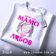 Футболка мама то любов, біла, зображена зверху, на фоні світло-сірого кольору, принт з текстом "Дякую тобі, МАМО" у яскраво-рожевому кольорі, з малюнком матері і дитини, на футболці також є етикетка.