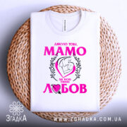 Футболка мама то любов, біла, зображена зверху, на фоні плетеного кошика, принт містить текст "Дякую тобі, МАМО" у яскраво-рожевому кольорі, з малюнком матері і дитини в серці.