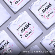 Футболка мама у світі, біла, зображена зверху на столі, фон світло-сірий, кілька футболок розкладені поруч, принт чорний з рожевими елементами та сердечками, текст говорить 'найкраща мама у світі'.