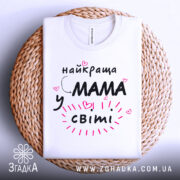 Футболка мама у світі, біла, зображена зверху на плетеній основі, фон білий, принт чорний з рожевими елементами та сердечками, текст говорить 'найкраща мама у світі'.