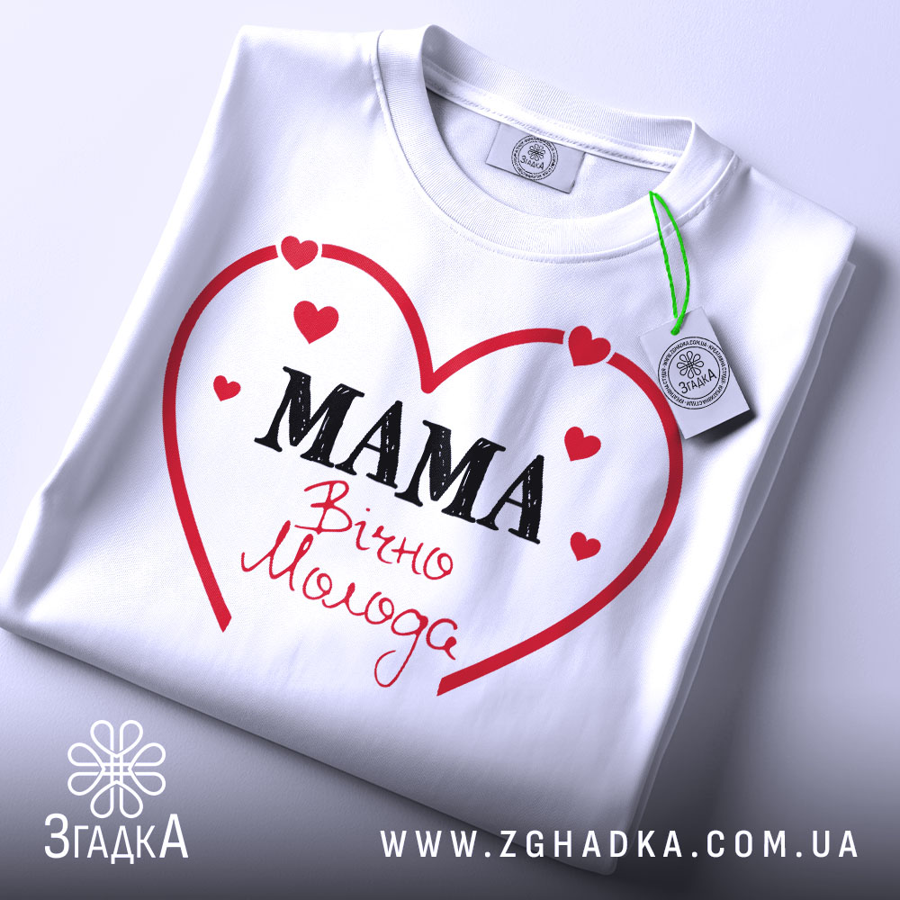 Футболка мама вічно молода, білого кольору, складена на поверхні, фон світло-сірий, принт у вигляді червоного серця з написом 'MAMA' чорним шрифтом та 'Вічно Молода' червоним шрифтом, з прикріпленою етикеткою.