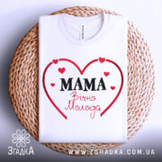 Футболка мама вічно молода, білого кольору, розкладена на плетеній основі, фон білий, принт у вигляді червоного серця з написом 'MAMA' чорним шрифтом та 'Вічно Молода' червоним шрифтом.