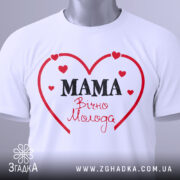 Футболка мама вічно молода, білого кольору, складена на поверхні, фон світло-сірий, принт у вигляді червоного серця з написом 'MAMA' чорним шрифтом та 'Вічно Молода' червоним шрифтом, з прикріпленою етикеткою.