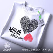 Футболка мама завжди поруч, білого кольору, складена на поверхні, фон світло-сірий, принт з сердець з відбитками пальців: чорне та червоне, текст чорного кольору, на комірці видна етикетка.