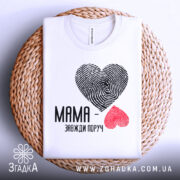 Футболка мама завжди поруч, білого кольору, складена на плетеній кошику, фон світло-сірий, принт з сердець з відбитками пальців: чорне та червоне, текст чорного кольору, видно округлий комірець.