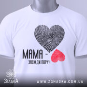 Футболка мама завжди поруч, білого кольору, складена на поверхні, фон світло-сірий, принт з сердець з відбитками пальців: чорне та червоне, текст чорного кольору, на комірці видна етикетка.