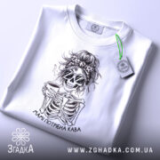 Футболка мамі потрібна кава, біла, складена на столі, фон світло-сірий, принт з черепом у сонцезахисних окулярах, що тримає чашку кави, з написом 'Мамі потрібна кава', на етикетці видно логотип бренду.