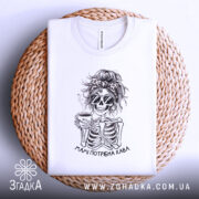 Футболка мамі потрібна кава, біла, складена на плетеному кошику, фон світло-сірий, принт з черепом у сонцезахисних окулярах, що тримає чашку кави, з написом 'Мамі потрібна кава', видно етикетку з логотипом бренду.