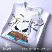 Футболка мамин день, біла, зображена з верхнього ракурсу, на фоні світло-сірого кольору, принт з двома птахами, що кричать 'MOM!', з зеленим ярликом.