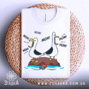 Футболка мамин день, біла, зображена зверху на плетеній основі, фон світло-сірий, принт з двома птахами, що кричать 'MOM!' та рибкою внизу.