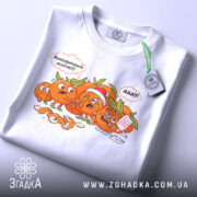 Футболка мандариновий маніяк, біла, складена на столі, з принтом мандаринів, що спілкуються, з етикеткою на зеленому шнурку, на фоні світлого кольору.