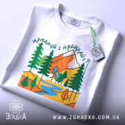 Футболка мандруй гори, біла, складена на поверхні, фон світло-сірий, принт з горами, деревами та річкою, етикетка з зеленим шнурком.