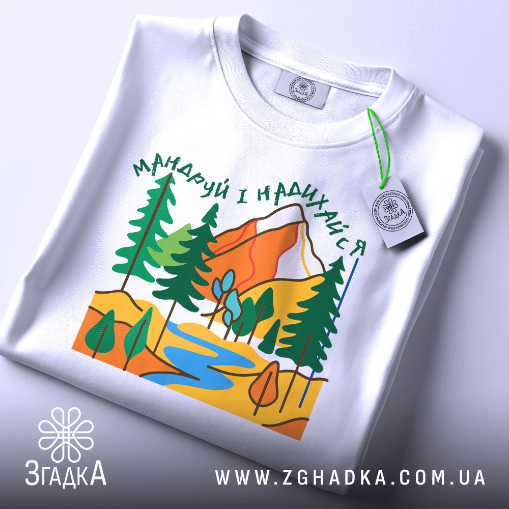 Футболка мандруй гори, біла, складена на поверхні, фон світло-сірий, принт з горами, деревами та річкою, етикетка з зеленим шнурком.