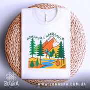 Футболка мандруй гори, біла, складена на плетеній кошику, фон білий, принт з горами, деревами та річкою, вид зверху.