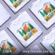 Футболка мандруй гори, біла, зображена на манекені, фон світло-сірий, принт з горами, деревами та річкою в яскравих кольорах.