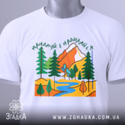 Футболка мандруй гори, біла, складена на поверхні, фон світло-сірий, принт з горами, деревами та річкою, етикетка з зеленим шнурком.