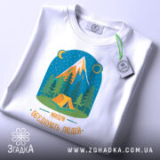 Футболка мандри обʼєднують білого кольору, складена на поверхні, фон світлий. Принт зображає гори, ліс і намет, кольори яскраві, текст 'Мандри обʼєднують людей' розташований внизу. На футболці є етикетка з зеленим шнурком.