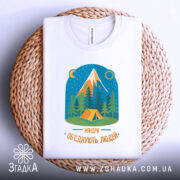 Футболка мандри обʼєднують білого кольору, складена на плетеній кошику, фон світлий. Принт містить малюнок гір, лісу та намету, кольори яскраві, текст 'Мандри обʼєднують людей' розташований внизу.