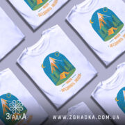 Футболка мандри обʼєднують білого кольору, зображена на манекені з блискучою поверхнею, фон нейтральний сірий. Принт містить малюнок гір, лісу та намету, виконаний у яскравих кольорах з текстом 'Мандри обʼєднують людей'.