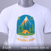 Футболка мандри обʼєднують білого кольору, складена на поверхні, фон світлий. Принт зображає гори, ліс і намет, кольори яскраві, текст 'Мандри обʼєднують людей' розташований внизу. На футболці є етикетка з зеленим шнурком.