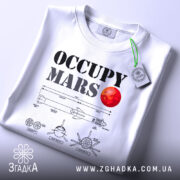 Футболка Марс колонізує біла, складена на поверхні, з принтом 'OCCUPY MARS' та червоною планетою, з зеленим ярликом, на фоні світло-сірого градієнту.
