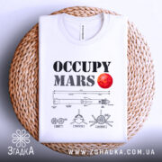 Футболка Марс колонізує біла, складена на плетеній кошику, з принтом 'OCCUPY MARS' та червоною планетою, на фоні нейтрального кольору.