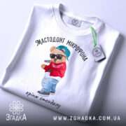 Футболка мастодонт мікрофона білого кольору, зображена зверху, на плоскій поверхні, фон світлий, принт з ведмедиком у червоній куртці та синій кепці, що тримає мікрофон, текст під принтом: "кринж стендапу", на комірці видна етикетка.