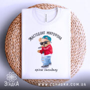 Футболка мастодонт мікрофона білого кольору, зображена зверху на плоскій поверхні з плетеного матеріалу, фон світлий, принт з ведмедиком у червоній куртці та синій кепці, що тримає мікрофон, текст під принтом: "кринж стендапу".