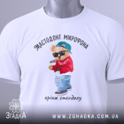 Футболка мастодонт мікрофона білого кольору, зображена зверху, на плоскій поверхні, фон світлий, принт з ведмедиком у червоній куртці та синій кепці, що тримає мікрофон, текст під принтом: "кринж стендапу", на комірці видна етикетка.