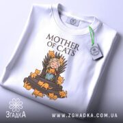 Футболка мати котів білого кольору, зображена в складеному вигляді на світлому фоні, принт з малюнком дівчини на троні з котами, напис 'MOTHER OF CATS' зверху, на футболці є етикетка з зеленим шнурком.