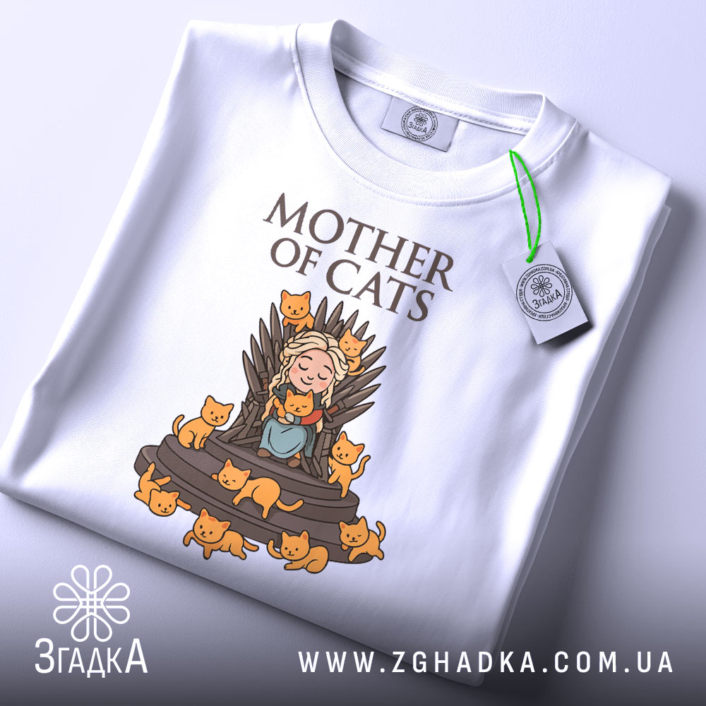 Футболка мати котів білого кольору, зображена в складеному вигляді на світлому фоні, принт з малюнком дівчини на троні з котами, напис 'MOTHER OF CATS' зверху, на футболці є етикетка з зеленим шнурком.