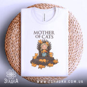 Футболка мати котів білого кольору, зображена в складеному вигляді на плетеній кошику, фон білий, принт з малюнком дівчини на троні з котами, напис 'MOTHER OF CATS' зверху.