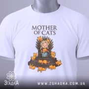 Футболка мати котів білого кольору, зображена в складеному вигляді на світлому фоні, принт з малюнком дівчини на троні з котами, напис 'MOTHER OF CATS' зверху, на футболці є етикетка з зеленим шнурком.