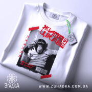 Футболка мавпа життя цікаве. Біла футболка, складена на столі, з принтом чорного мавпи, що курить. На фоні світло-сірого кольору, з зеленим ярликом.