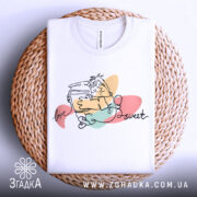 Футболка медовий настрій, біла, зображена зверху на плетеній основі, фон світлий, принт з малюнком персонажа, що тримає банку з медом, з написом 'love sweet', видна етикетка на комірці.