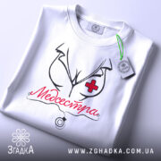 Футболка медсестра білого кольору, складена на столі, з видимим етикеткою, фон світло-сірий, принт з малюнком медсестри та написом 'Медсестра' червоного кольору.