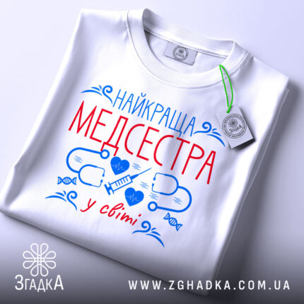 Футболка медсестра білого кольору, складена на поверхні, фон світлий, принт з червоним текстом "НАЙКРАЩА МЕДСЕСТРА" та синіми медичними символами, на комірці видна етикетка.