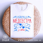 Футболка медсестра білого кольору, складена на плетеній основі, фон світлий, принт з червоним текстом "НАЙКРАЩА МЕДСЕСТРА" та синіми медичними символами.