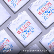 Футболка медсестра білого кольору, зображена на манекені, фон світло-сірий, принт містить червоний текст "НАЙКРАЩА МЕДСЕСТРА" та сині медичні символи.