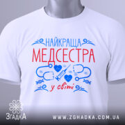 Футболка медсестра білого кольору, складена на поверхні, фон світлий, принт з червоним текстом "НАЙКРАЩА МЕДСЕСТРА" та синіми медичними символами, на комірці видна етикетка.