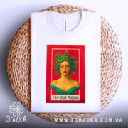 Футболка медуза я отрута, біла футболка, складена на плетеній кошику, з принтом жінки з зеленим волоссям на червоному фоні. Видно етикетку на горловині, фон світлий.