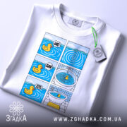 Футболка Мем Качечка, біла, лежить на столі, фон світлий, принт з шести коміксів з качечкою, яскраві кольори, на комірці є етикетка.