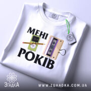 Футболка мені років. Біла футболка, складена на столі, з принтом, що містить малюнки касети та персонажа з олівцем, з етикеткою на зеленому шнурку, фон світло-сірий.