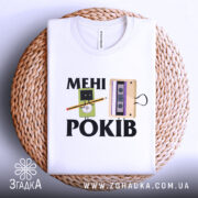 Футболка мені років. Біла футболка, складена на плетеній кошику, з принтом, що містить малюнки касети та персонажа з олівцем, фон світло-сірий.