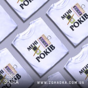 Футболка мені років. Біла футболка на манекені, з принтом, що містить малюнки касети та персонажа з олівцем, фон світло-сірий.