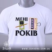 Футболка мені років. Біла футболка, складена на столі, з принтом, що містить малюнки касети та персонажа з олівцем, з етикеткою на зеленому шнурку, фон світло-сірий.