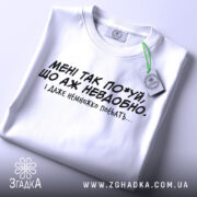 Футболка мені так по*уй, біла футболка, складена на столі, з чорним принтом, текст на грудях: 'МЕНІ ТАК ПО*УЙ, ЩО АЖ НЕВДОБНО. І ДАЖЕ НЕМНОЖКО ПОБЛЯТЬ...'. На футболці є етикетка.