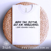 Футболка мені так по*уй, біла футболка, складена на плетеній кошику, з чорним принтом, текст на грудях: 'МЕНІ ТАК ПО*УЙ, ЩО АЖ НЕВДОБНО. І ДАЖЕ НЕМНОЖКО ПОБЛЯТЬ...'. Фон світлий.