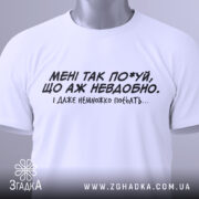 Футболка мені так по*уй, біла футболка, складена на столі, з чорним принтом, текст на грудях: 'МЕНІ ТАК ПО*УЙ, ЩО АЖ НЕВДОБНО. І ДАЖЕ НЕМНОЖКО ПОБЛЯТЬ...'. На футболці є етикетка.