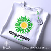 Футболка ментальне здоров`я білого кольору, зображена зверху, на плоскій поверхні. Принт з жовтим сонцем та зеленими листками, текст чорного кольору: 'МЕНТАЛЬНЕ ЗДОРОВ'Я Ти як?'. На футболці підвішена етикетка.